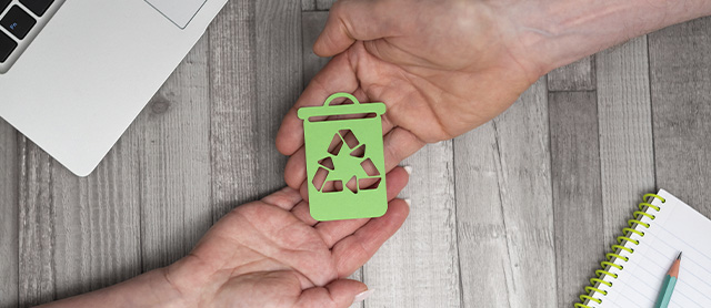 duas mãos segurando um lixo reciclável feito de papel verde em cima de uma mesa de escritório