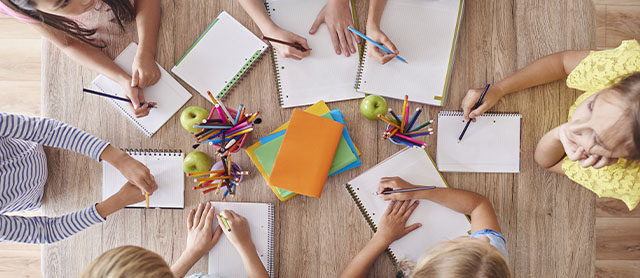 crianças sentadas ao redor de uma mesa conversando e usando materiais escolares para desenhar e estimular sua criatividade